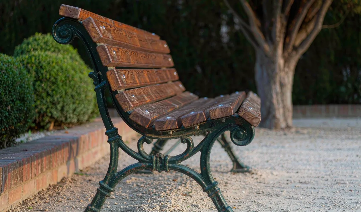 Cast iron bench ends painted dark green holding wooden slats bolted through the ironwork, on a gravel pad beside a low brick wall
